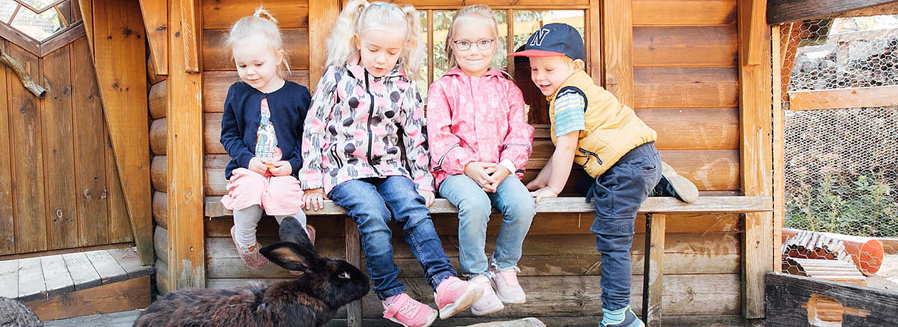 Vier kleine Kinder auf einer Holzbank. Vor ihnen sitzt ein Hase.