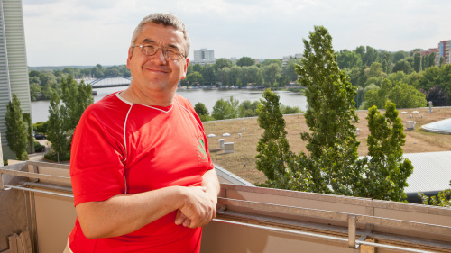 Ein Mann steht auf seinem Balkon und blickt stolz und froh in die Kamera. Im Hintergrund ist der Ausblick auf Wiese, Wald und See zu sehen.