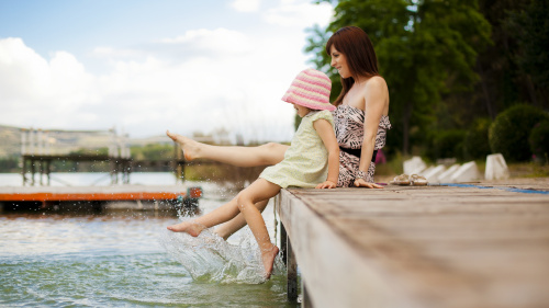 Eine Mutter sitzt mit ihrer Tochter am Wasser und sie lassen die Füße ins Wasser baumeln. 