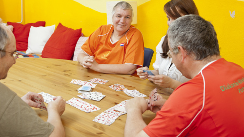 Männer und Frauen sitzen an einem Tisch und spielen Karten. Eine Frau in einem leuchtenden orangenen T-Shirt schaut in die Kamera und lächelt.