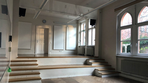 Blick zur Tür in einem großen hellen Raum mit großen Fenster, ein paar Treppen und dem Parkettboden