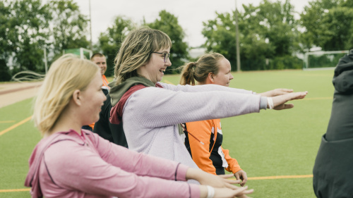 Gruppe macht gemeinsam Dehnübungen mit ausgestreckten Armen auf Sportplatz