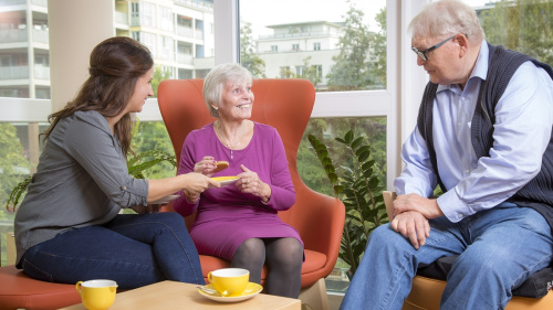 Eine Seniorin sitzt mit zwei Personen zusammen und genießt fröhlich Tee und einen Keks.
