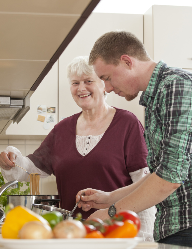 Ein junger Man udn eine ältere Dame stehen in der Küche an der Arbeitsplatte und bereiten gemeinsam ein essen vor. Die Dame lächelt in die Kamera.