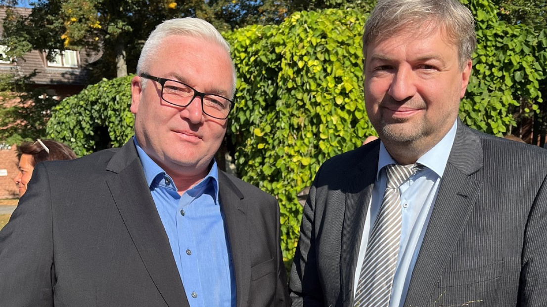 Zwei Herren stehen neben einander und schauen lächelnd in die Kamera. Ein Herr hat eine Urkunde und eine Sonnenblume in den Händen. Der andere Herr hält einen Briefumschlag in seiner Hand.