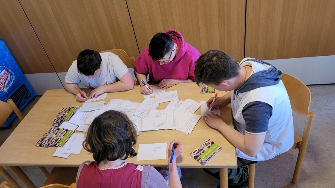 Eine Gruppe von Menschen sitzt gebeugt über einem runden Tisch. Alle schreiben konzentriert auf DIN A5 große Karteikarten.