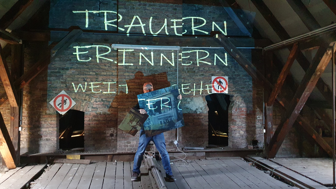 Ein älterer Herr mit Kappe und Brille steht breitbeinig in einem staubigen Dachstuhl eines alten Backstein-Hauses. Er hält groß ausgedruckte Fotos hoch und verdeckt damit halb sein Gesicht. Eine Beamer-Projektion strahlt auf ihn und die Wand hinter ihm die Schrift "Trauern, erinnern, weitergehen"
