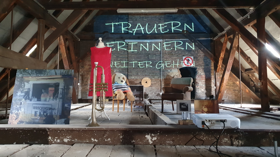 Auf einem verstaubten Dachstuhl stehen verschiedene Gegenstände: Eine Trompete, ein rotes Kleid, ein großer Teddybär, der auf einem Stuhl sitzt, ein brauner Sessel, große Fotodrucke. Ein Beamer strahlt die Schrift "Trauern, Erinnern, Weitergehen" auf die Wand im Hintergrund