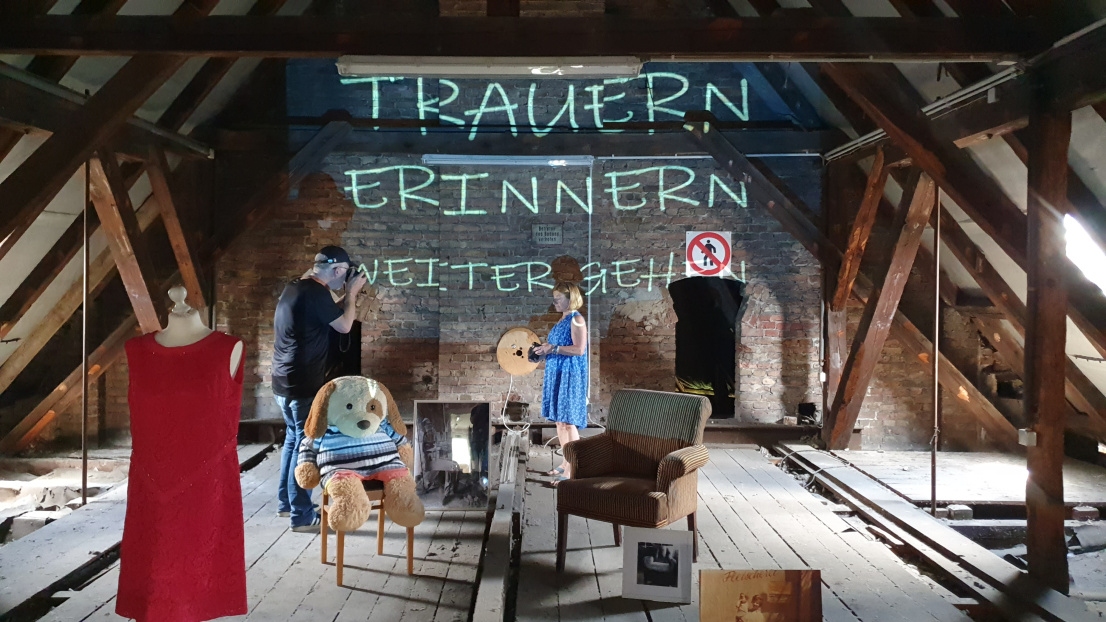 Auf einem verstaubten Dachstuhl stehen verschiedene Gegenstände: Eine Trompete, ein rotes Kleid, ein großer Teddybär, der auf einem Stuhl sitzt, ein brauner Sessel, große Fotodrucke. Ein Beamer strahlt die Schrift "Trauern, Erinnern, Weitergehen" auf die Wand im Hintergrund. Zwei Menschen im Hintergrund machen Fotos