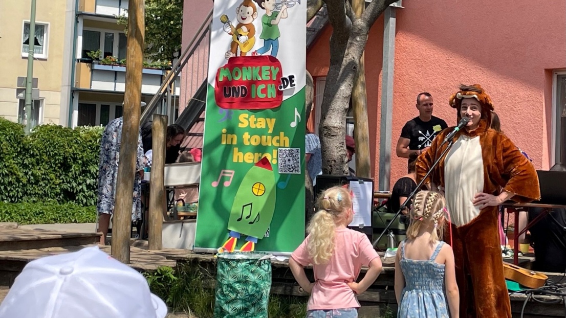 Vor einem rosa zweistöckigen Gebäude steht ein Baum. Davor hängt ein Banner mit der Aufschrift "Monkey und ich.de Stay in touch here". Daneben steht eine verkleidete Frau im Affen-Kostüm, die in ein Mikrofon spricht. Um sie herum stehen Kinder, im Hintergrund Erwachsene.