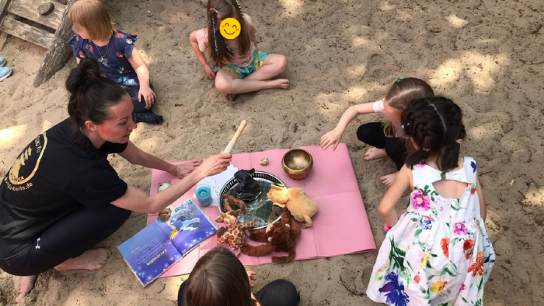 Eine Frau sitzt mit Kindern auf Sand neben einem Klettergerüst. Sie hat eine Decke ausgebreitet, auf der eine Klangschale und Kuscheltiere liegen.
