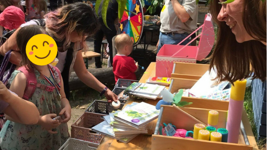 Auf einem langen Holztisch im Freien liegen Bücher und Kosten voller Spielzeug und Bastelmaterialien. Eine Frau und ein Mädchen lächeln sich an, im Hintergrund sind kleine und große Menschen sowie überdachte Bierbänke zu sehen. Die Sonne strahlt warm.