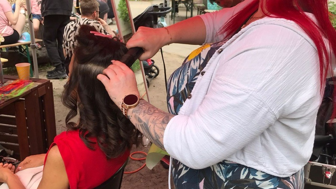 Unter einem Zelt im Freien steht eine Frau mit langen roten Haaren und Sonnenbrille. Sie macht einer jungen Frau auf einem Stuhl vor ihr eine hübsche Frisur mit einem Lockenstab. Im Hintergrund sind viele Menschen und Wimpelketten zu sehen.