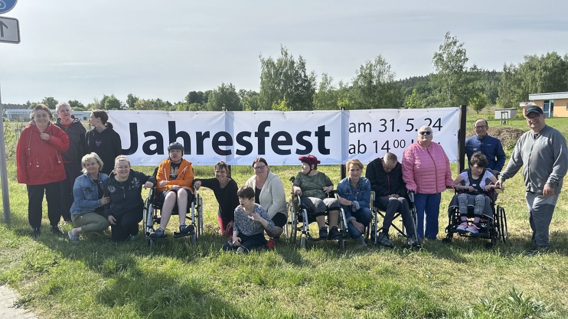 Eine bunte Gruppe von Menschen mit und ohne Beeinträchtigung steht auf einer weiten Wiese vor einem großen, weißen Plakat mit der Aufschrift "Jahresfest"