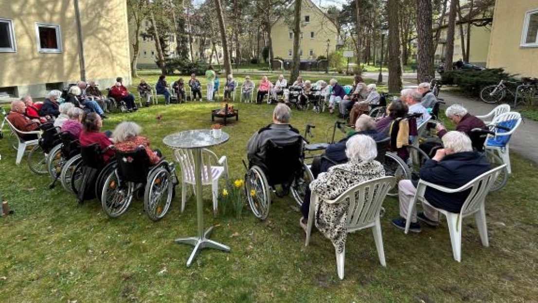 Viele Senioren und Seniorinnen sitzen in einem großen Kreis auf einer Wiese. Manche von ihnen sitzen in Rollstühlen, manche auf Stühlen oder Rollatoren.