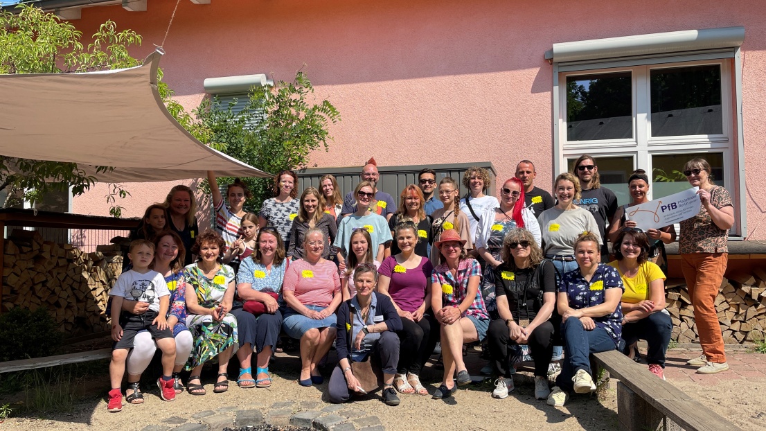 Eine Gruppe von circa 30 erwachsenen Menschen steht lächelnd in der Sonne vor einem einstöckigen rosa Gebäude. Sie tragen alle gelbe Namensschilder auf der Brust. Teilweise sitzen Kinder auf dem Schoß.