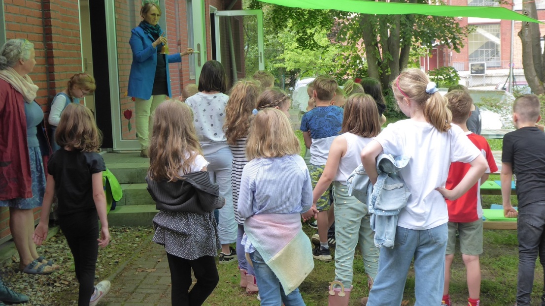 Viele Kinder stehen draußen unter grünen Sonnensegeln um den Eingang eines Gebäudes und hören einer Frau zu. Die Frau hält die Eingangstür auf und spricht in ein Mikrofon.