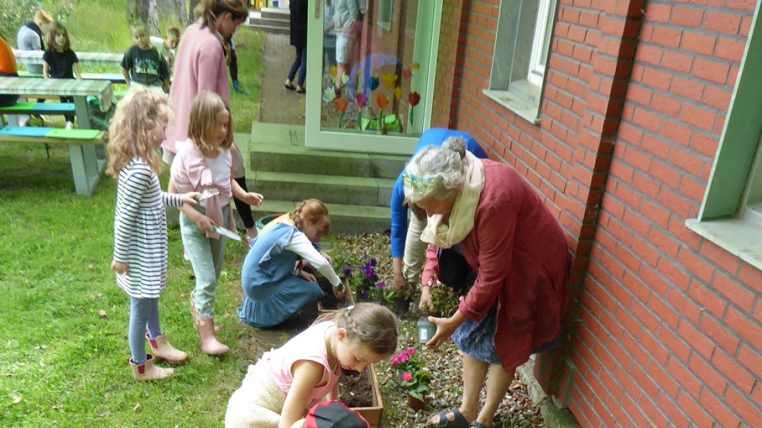 Einige Kinder bepflanzen zusammen mit Erwachsenen Blumenkästen auf dem Boden. Sie halten Schüppen in den Händen und es liegen bunte Beetpflanzen-Setzlinge rundeherum zur Bepflanzung bereit.