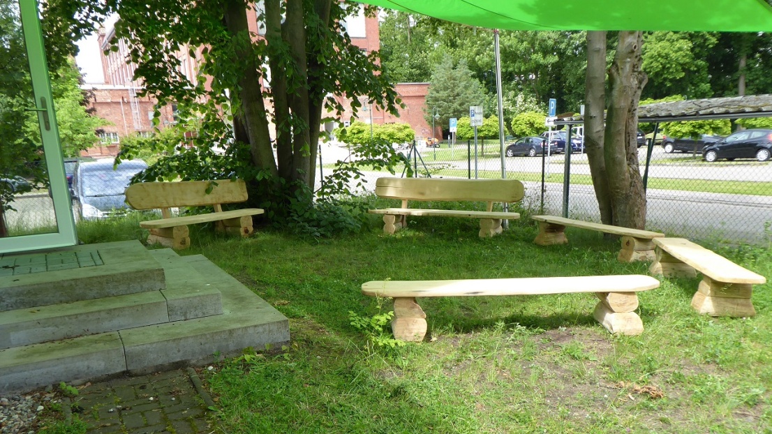 Auf einer grünen Wiese und unter grünen Sonnensegeln stehen im Halbschatten helle, neue Holzbänke.