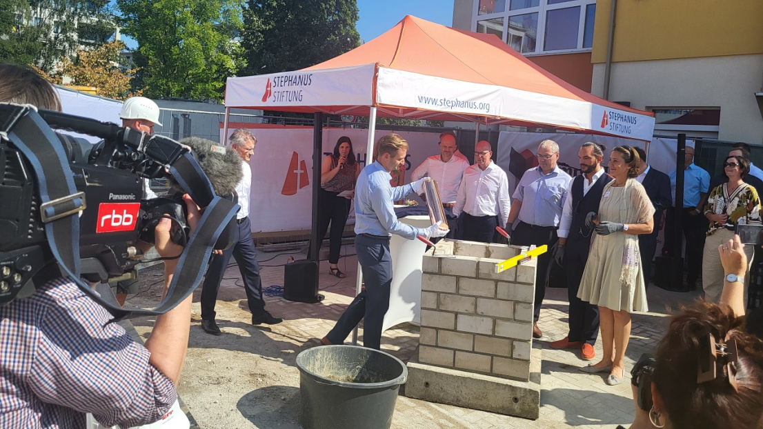 Bei sommerlich-warmen Wetter stehen einige schick gekleidete Menschen (darunter der Vorstand der Stephanus-Stiftung) um einen kleinen Sockel, der aus einzelnen Formsteinen gemauert ist. Ein Eimer Zement steht daneben. Im Vordergrund des Bildes erkennt man eine Person, die eine große Filmkamera von "rbb" trägt und die Personen um den Sockel herum filmt. Im Hintergrund befindet sich ein oranges Festzelt mit der Aufschrift "Stephanus"