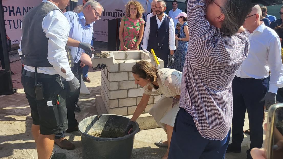 Bei sommerlich-warmen Wetter legt der Vorstand der Stephanus-Stiftung Hand an: Frau Dr. Ellen Ueberschär und Harald Thiel befestigen mit einer Schaufel Zement einen Formstein auf einem ersten Grundbau-Sockel. Im Vordergrund des Bildes erkennt man eine Person, die eine große Filmkamera von "rbb" trägt und die Personen um den Sockel herum filmt. Im Hintergrund befindet sich ein oranges Festzelt mit der Aufschrift "Stephanus"