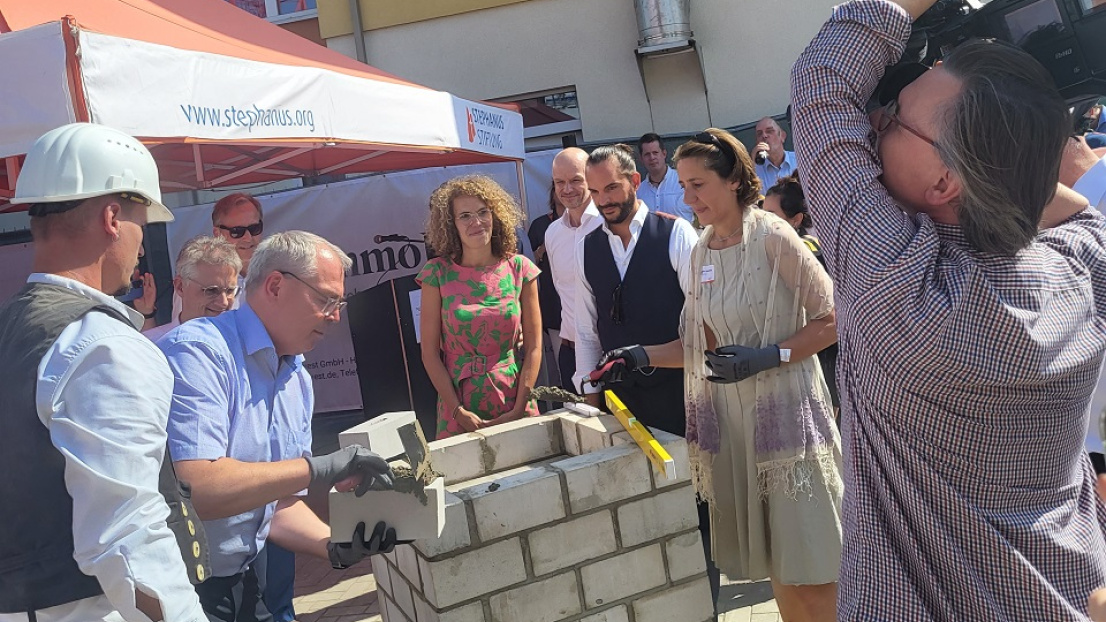Bei sommerlich-warmen Wetter legt der Vorstand der Stephanus-Stiftung Hand an: Frau Dr. Ellen Ueberschär und Harald Thiel befestigen mit einer Schaufel Zement einen Formstein auf einem ersten Grundbau-Sockel. Im Vordergrund des Bildes erkennt man eine Person, die eine große Filmkamera von "rbb" trägt und die Personen um den Sockel herum filmt. Im Hintergrund befindet sich ein oranges Festzelt mit der Aufschrift "Stephanus"