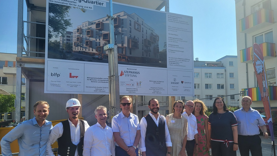 Bei sommerlich-warmen Wetter steht eine Gruppe von fröhlich lächelnden Menschen vor einem Baustellen-Plakat mit der Aufschrift "Schloßberg Quartier". Unter den Personen befindet sich der Vorstand der Stephanus-Stiftung: Frau Dr. Ellen Ueberschär und Harald Thiel.