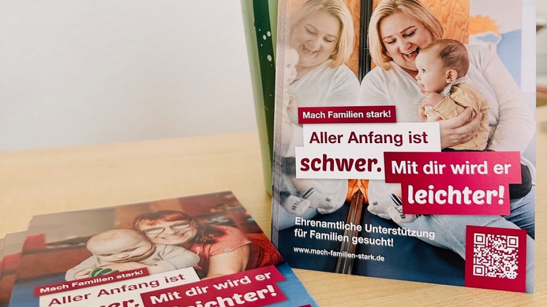 Eine Nahaufnahme von Flyern zum Thema "Ehrenamtliche Unterstützung für Familien": Eine Frau hält freudestrahlend ein Kleinkind im Arm. Darüber der Slogan "Aller Anfang ist schwer. Mit dir wird er leichter!"