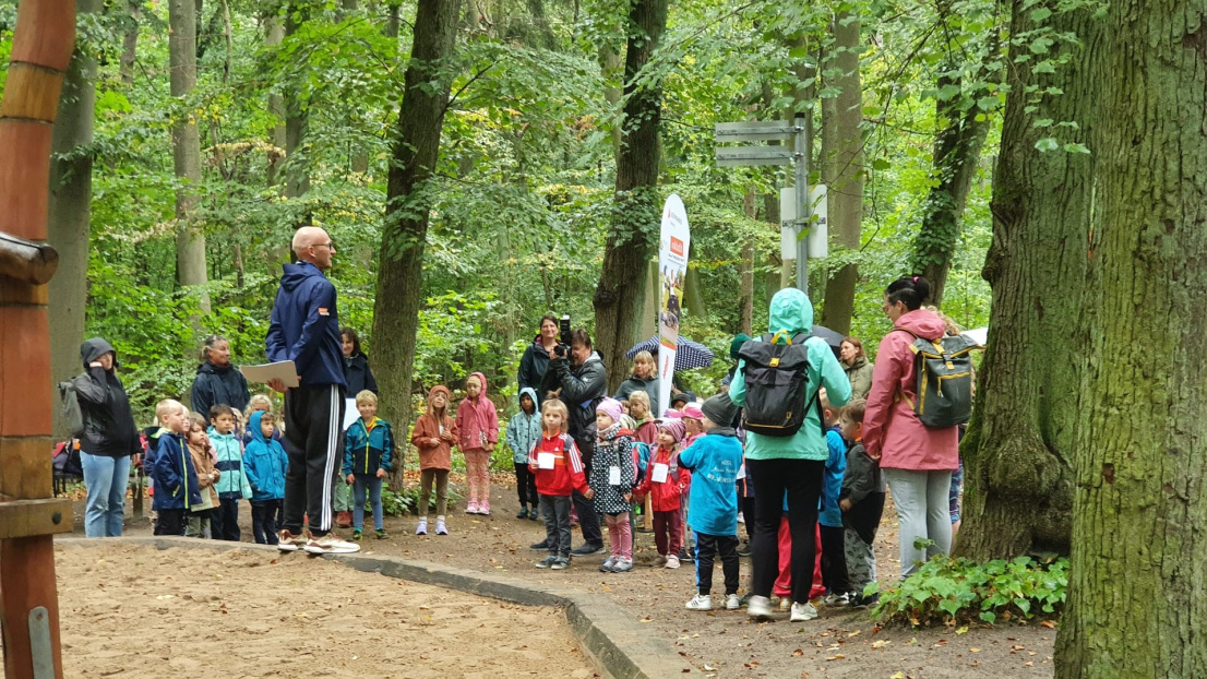 Eine Gruppe von Kindern und Eltern steht in Jacken gekleidet im Wald und hört gespannt einem sportlich gekleideten Herrn mit Glatze und Brille zu.