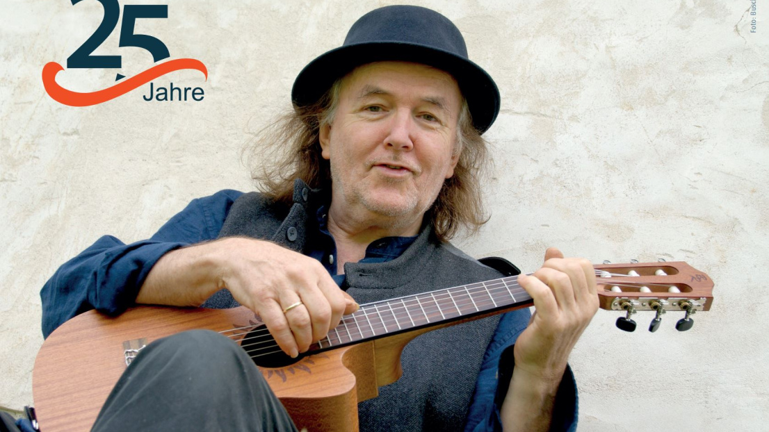 Ein Mann mit Hut und auf einer kleinen Gitarre spielend blickt freundlich in die Kamera