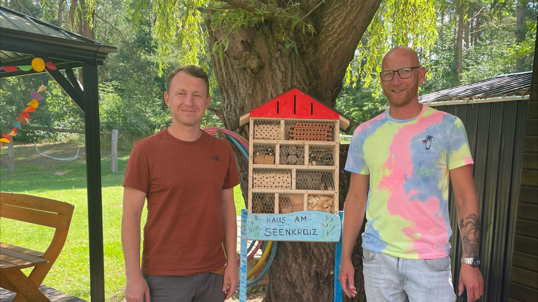 Zwei Männer stehen unter einem Baum und präsentieren stolz und fröhlich ein selbst gebautes Insektenhotel