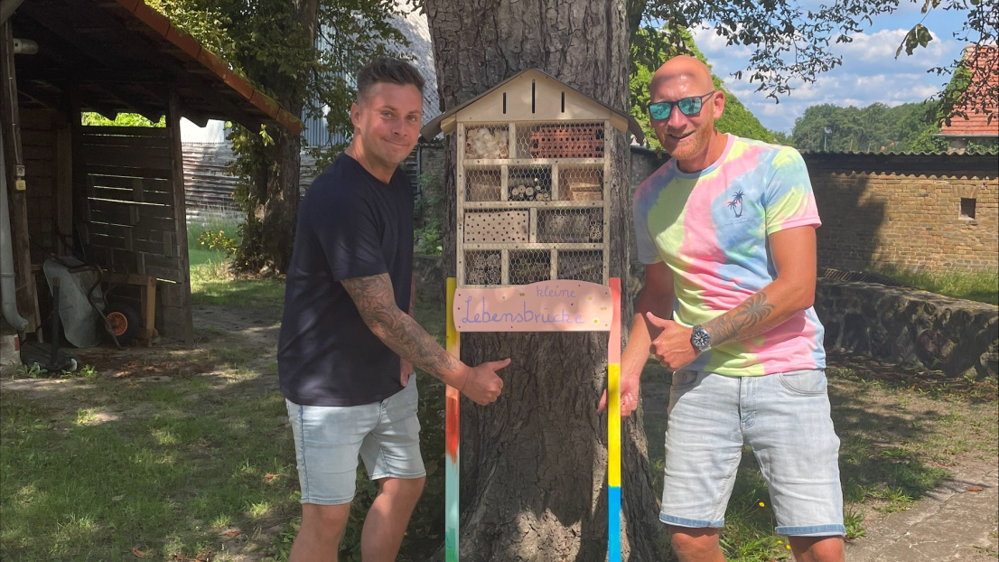 Zwei Männer stehen unter einem Baum und präsentieren stolz und fröhlich ein selbst gebautes Insektenhotel