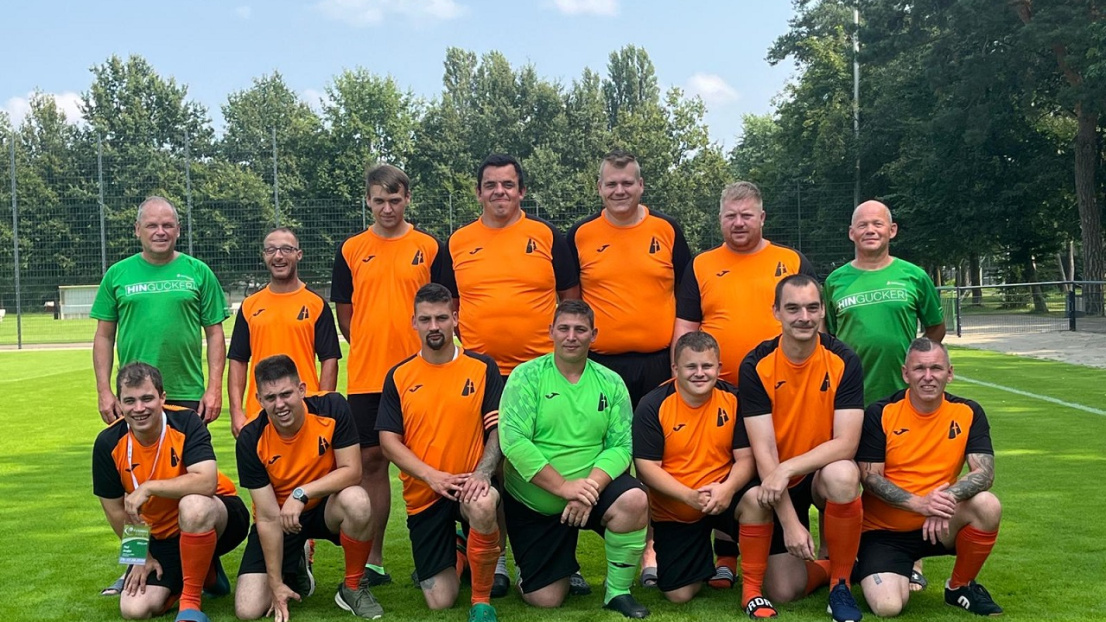 Eine fröhlich-motivierte Fußballmannschaft steht aufgereiht auf einem Fußballplatz. Sie tragen orange und grüne Trikots mit dem Stephanus-Logo