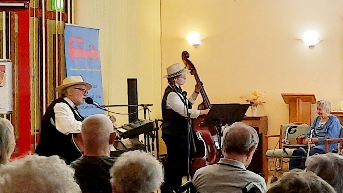 Zwei Musikanten spielen vor einem älteren Publikum in einem Saal. Beide tragen Hüte.
