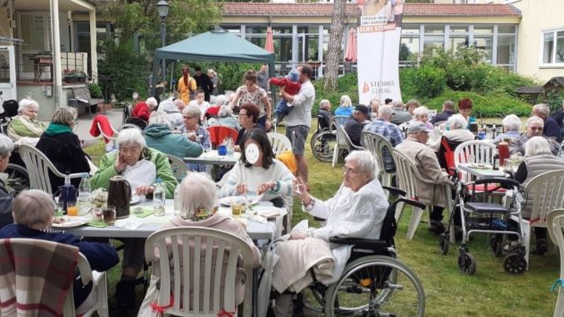 Auf einer Wiese sitzen viele Senioren und Seniorinnen auf weißen Plastikstühlen und gedeckten Kaffeetischen. Im Hintergrund ist ein gelbes Haus erkennbar.