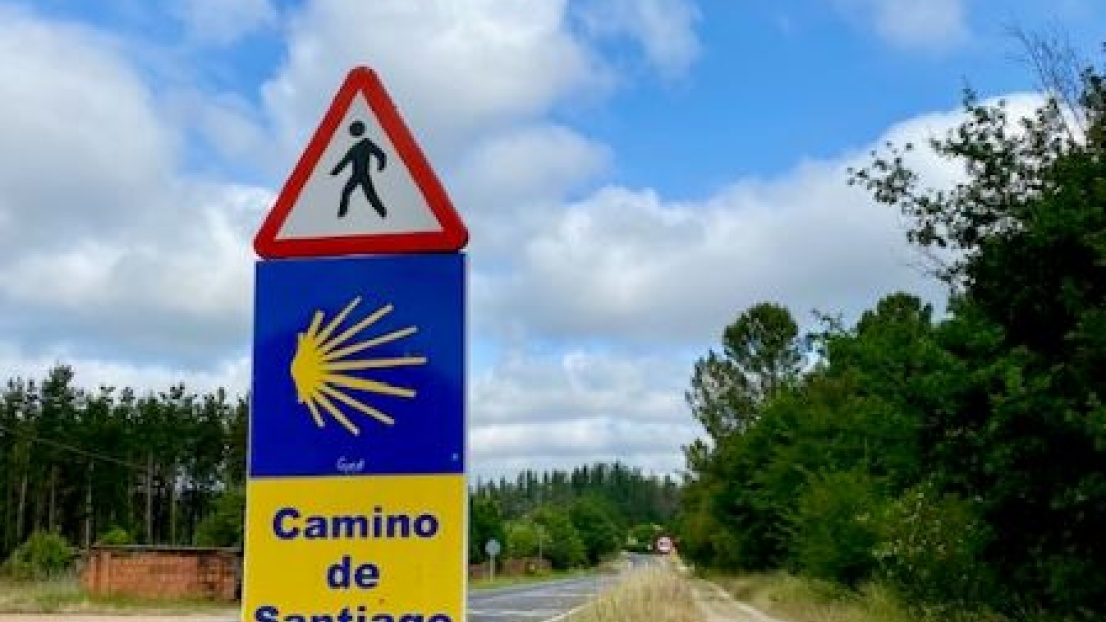 Eine Straße in sommerlicher Landschaft. Im Vordergrund steht ein Straßenschild mit dem Symbol des Jakobswegs und einem Fußgänger