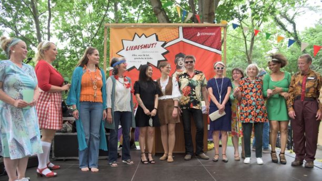 Auf einer kleinen Bühne im Freien steht eine Gruppe Menschen mit zum Teil lustigen, zum Teil schicken Kostümen. Sie lachen. Im Hintergrund hängen Wimpelketten und das Stephanus-Jahresmotto-Plakat mit der Aufschrift "Liebesmut!"