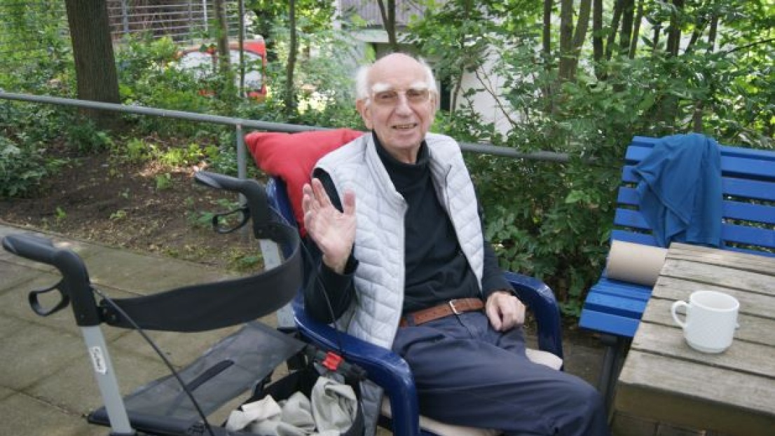 Ein älterer Herr lächelt winkend in die Kamera. Er sitzt mit einer Kaffeetasse an einem Tisch im Freien. Neben ihm steht ein Rollator.