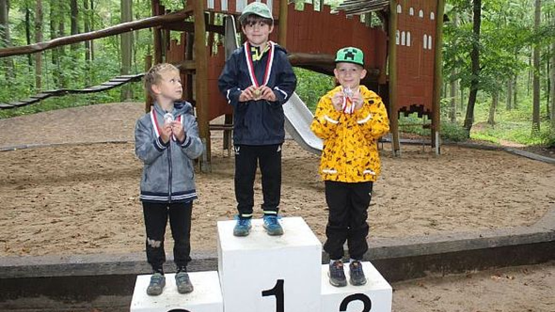 Drei Jungen stehen auf einem Siegerpodest im Wald auf einem 1., 2. und 3. Platz. Sie tragen jeweils eine Medaille. Im Hintergrund befindet sich ein Spielplatz.