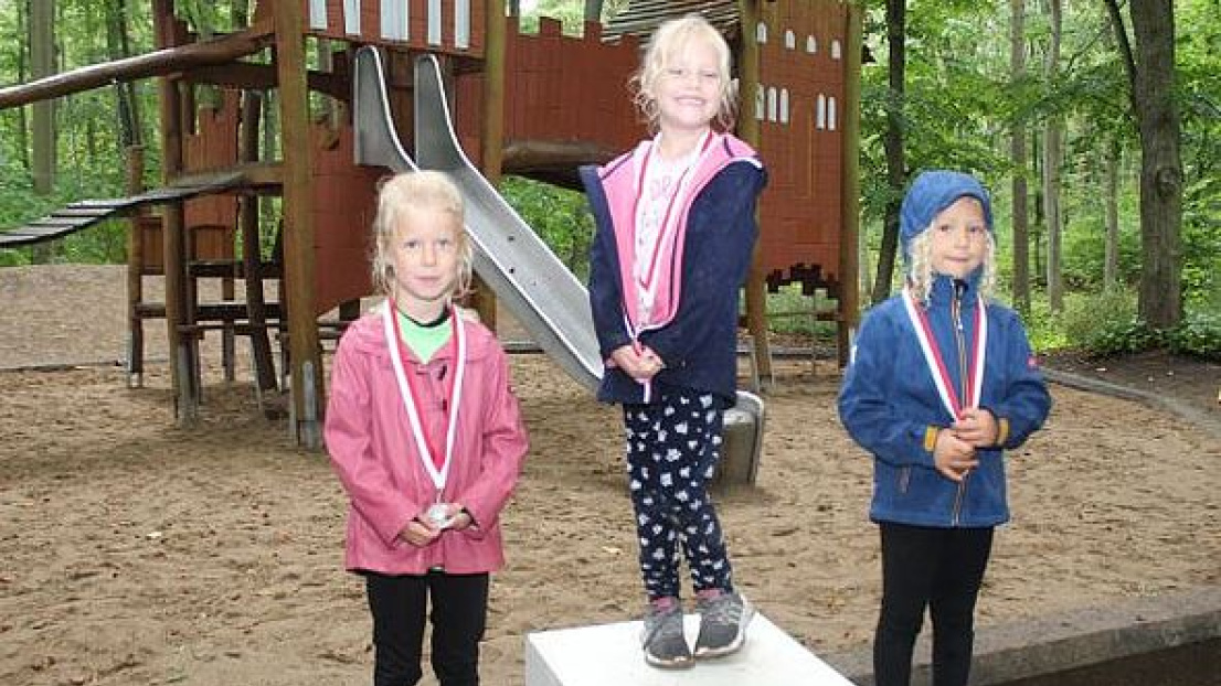 Drei Mädchen stehen auf einem Siegerpodest im Wald auf einem 1., 2. und 3. Platz. Sie tragen jeweils eine Medaille. Im Hintergrund befindet sich ein Spielplatz.