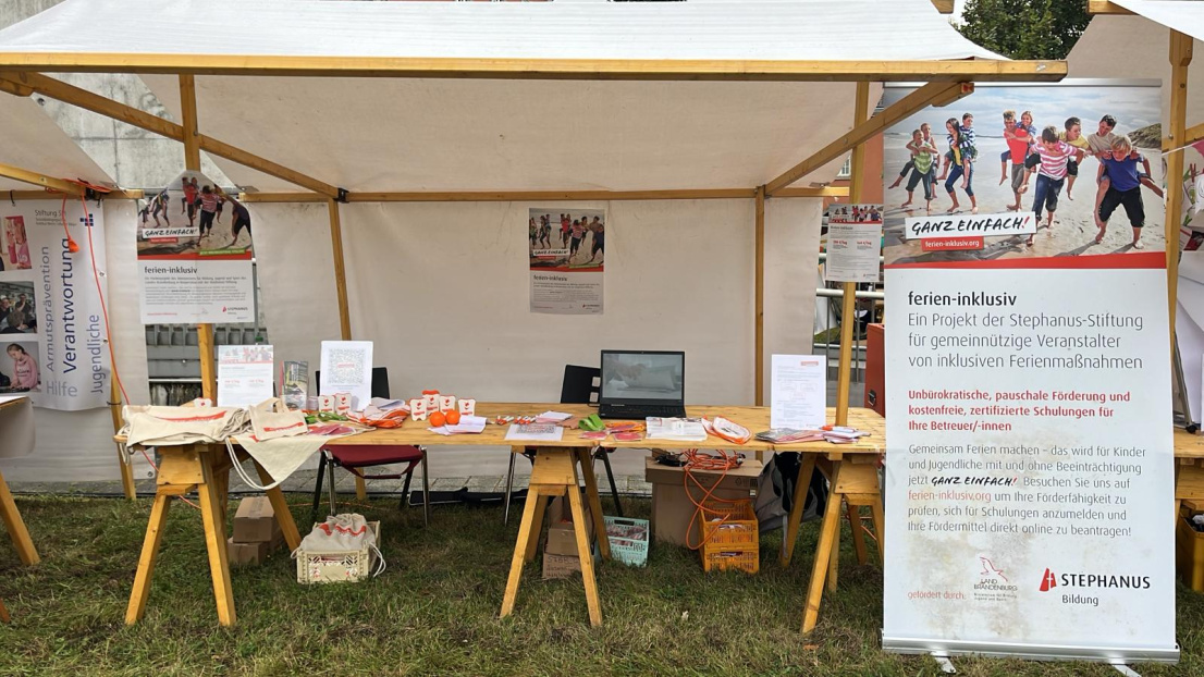 Ein Informationsstand auf einer Wiese, überdacht von einem weißen Leinen-Pavillon. Auf den Holztischen liegen Informationsmaterialien und Werbegeschenke bereit. Ein großer Banner steht daneben auf dem Boden und informiert über "ferien-inklusiv"