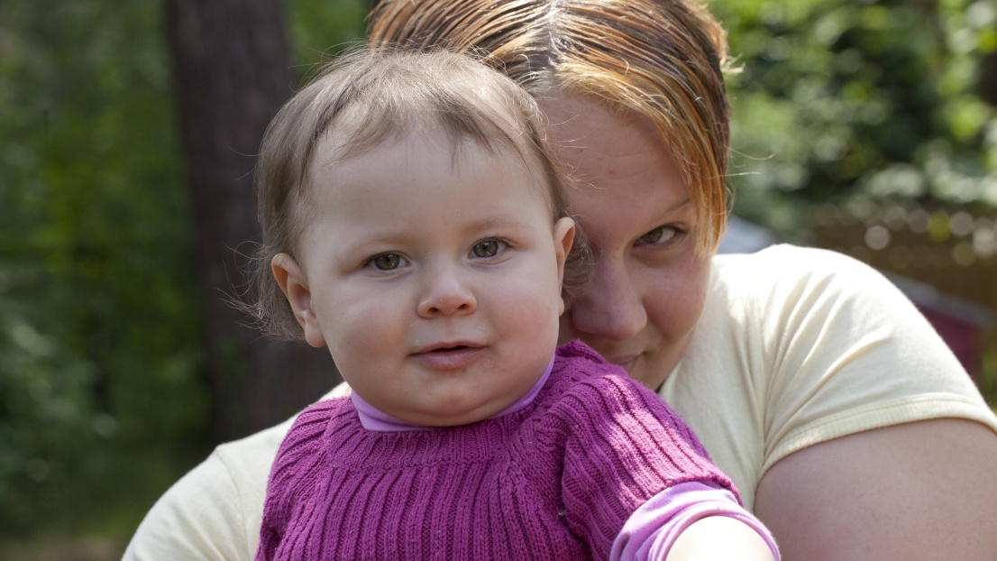 Eine junge Mutter hat ein Baby auf dem Schoß. Sie sitzen draußen, es ist Sommer. Die Mutter versteckt ihr Gesicht ein wenig hinter dem Baby, das Baby lächelt in die Kamera.