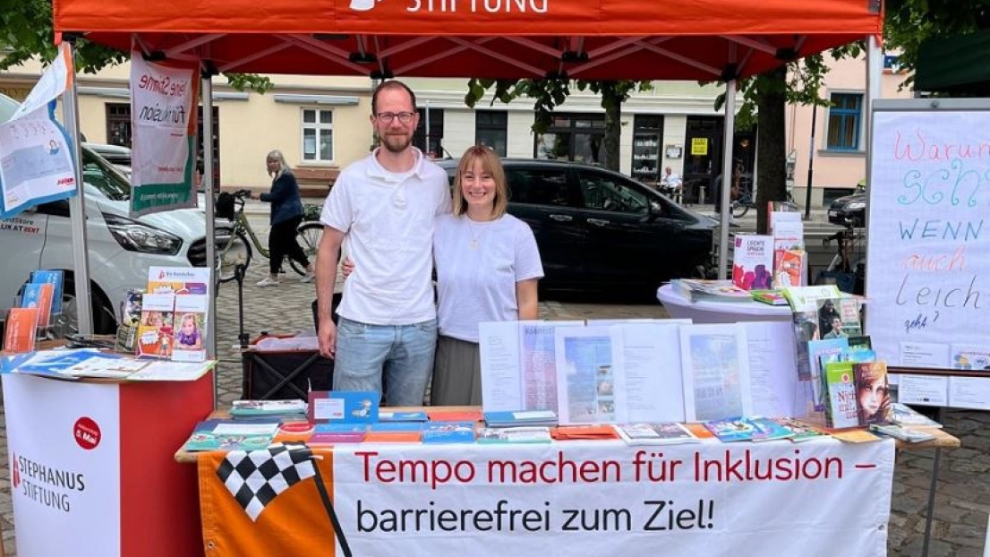Eine Frau und ein Mann stehen freundlich lächelnd Arm in Arm hinter einem überdachten Informationsstand auf einem Marktplatz. Auf dem Stand liegen viele verschiedene Flyer.