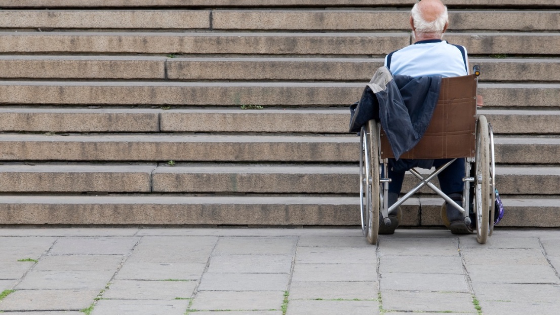 Ein Mann sitzt mit dem Rücken zur Kamera in einem Rollstuhl und schaut auf eine große Treppe vor sich