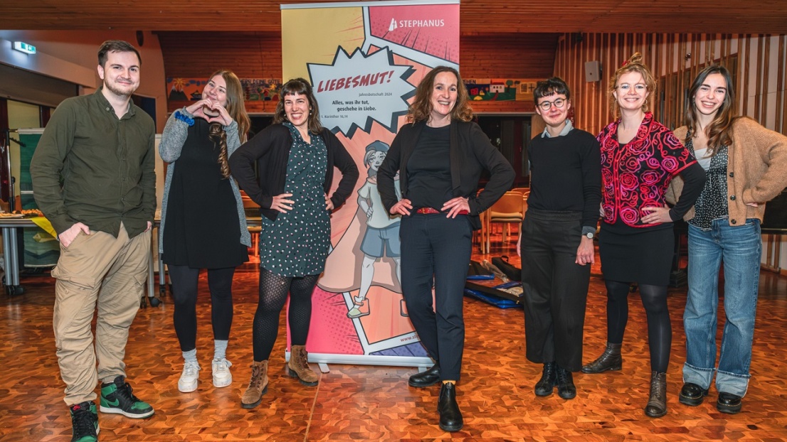 Sechs Frauen und ein Mann stehen fröhlich lächelnd in einem Saal vor einem Plakat mit dem Stephanus-Jahresmotto 2024 "Liebesmut!"