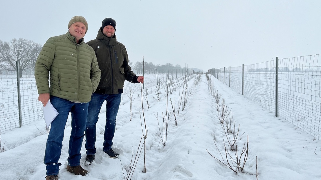 Zwei Männer stehen in einer einer weiß verschneiten Winterlandschaft auf einem Feld und lächeln in die Kamera
