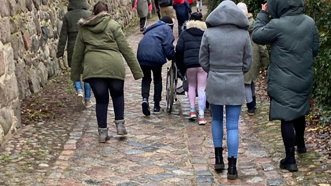 Kinder und Jugendliche mit und ohne Rollstuhl bewegen sich auf einem Pflasterweg an einer Stadtmauer entlang.