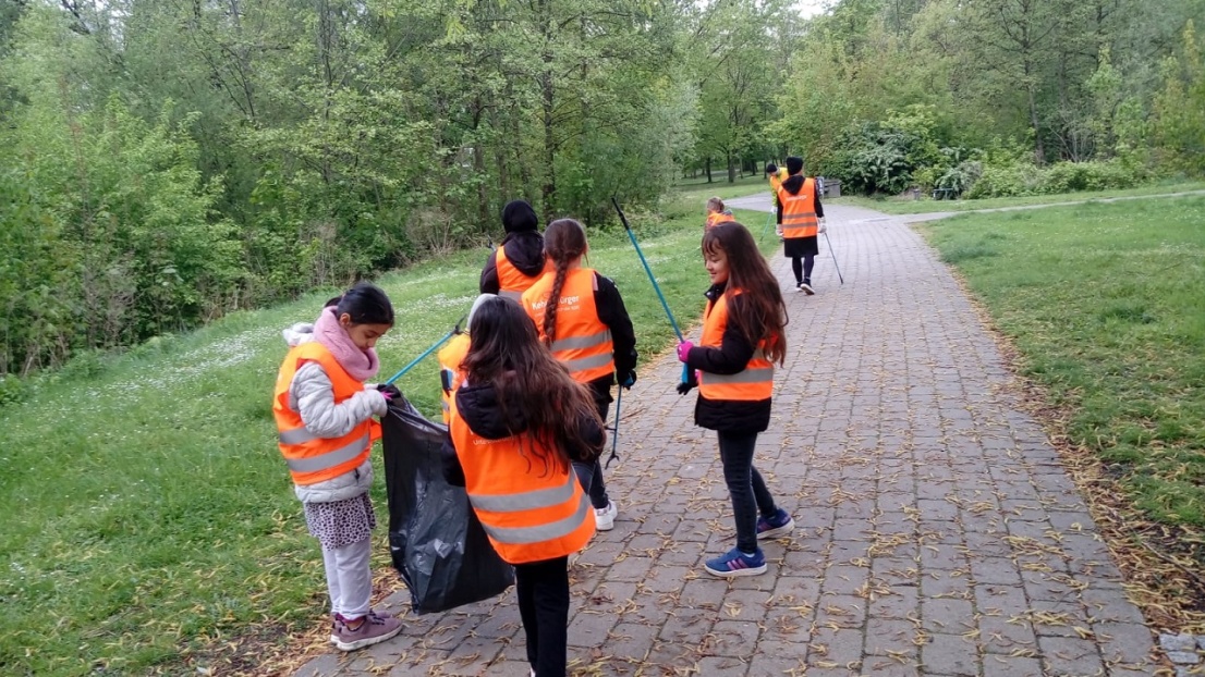 Kinder tragen orange Warnwesten und laufen mit Müllzangen und Müllbeuteln durch einen Park