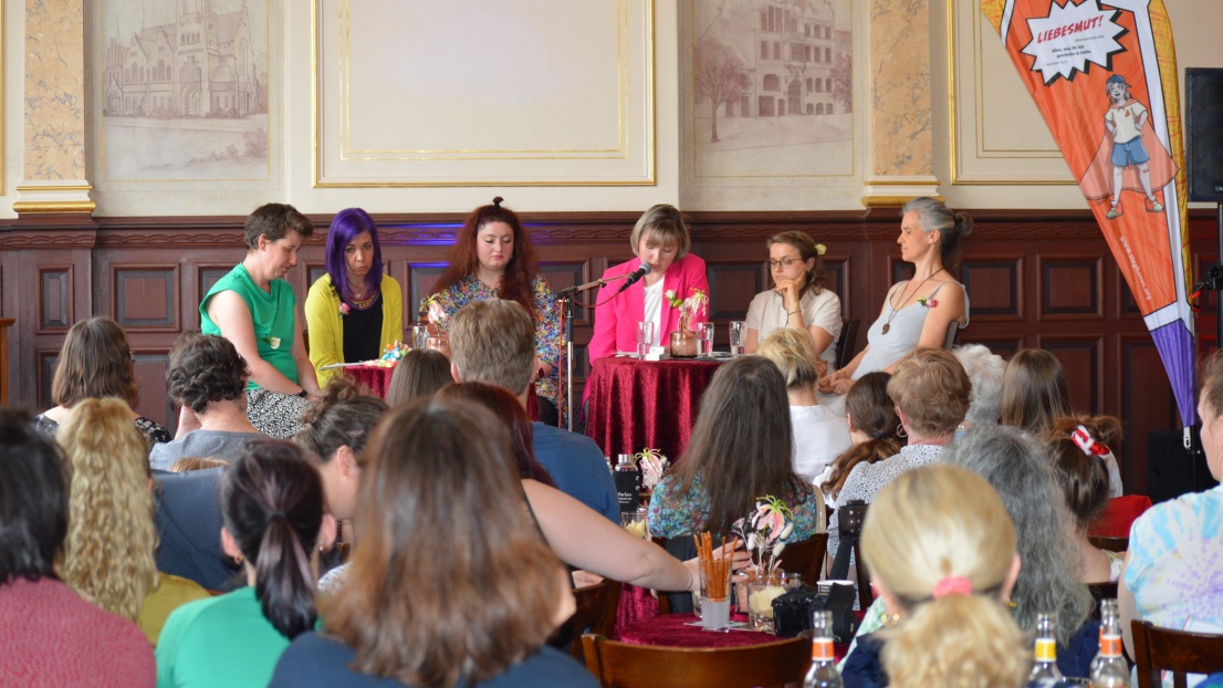 Ein Publikum in Rückenansicht schaut nach vorne auf eine Bühne, wo 6 Autorinnen sitzen und per Mikrofon aus dem Buch vorlesen