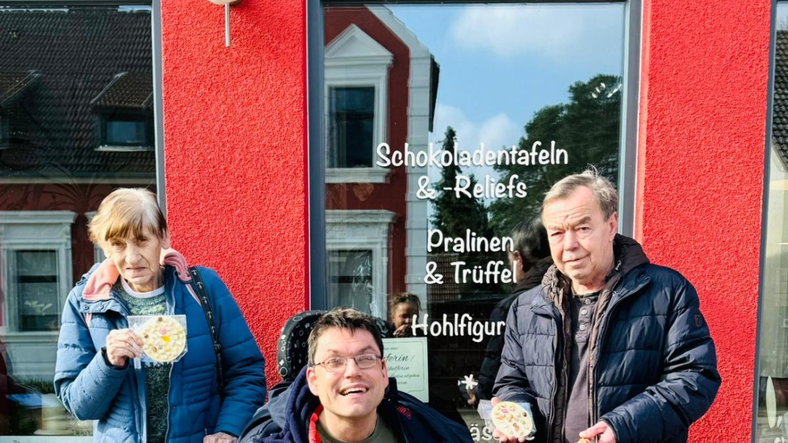 Drei Menschen, einer davon im Rollstuhl, stehen vor einem roten Haus und lächeln in die Kamera. Sie halten selbstgemachte Schokolade in der Hand.